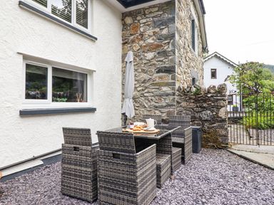 An outdoor seating area with a wicker table and chairs on a gravel surface next to a stone and white stucco building at Coed Derw Isaf in Betws-Y-Coed