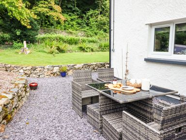 An outdoor patio area with a glass top table and wicker chairs on a stone ground near a white wall with a window at Coed Derw Isaf in Betws-Y-Coed