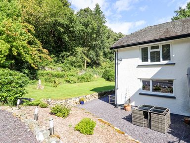 A garden area with shrubs and a lawn next to a white house with rattan outdoor furniture at Coed Derw Isaf in Betws-Y-Coed