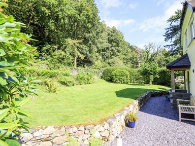 A garden with a green lawn and trees next to a stone wall and a house at Coed Derw Isaf in Betws-Y-Coed
