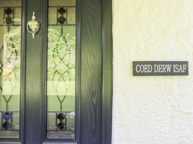 A black front door with glass panels and a brass knocker beside a white wall with a sign that reads Coed Derw Isaf at Betws Y Coed