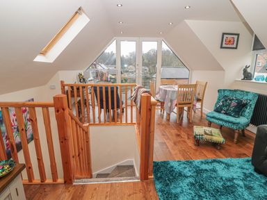 An attic living space with wooden railing stairs a dining table with four chairs a teal armchair and ottoman a blue rug and large windows at River Cottage in Morpeth