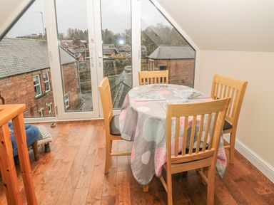 A dining area with a round table covered by a tablecloth and four wooden chairs near a glass door overlooking buildings at River Cottage in Morpeth