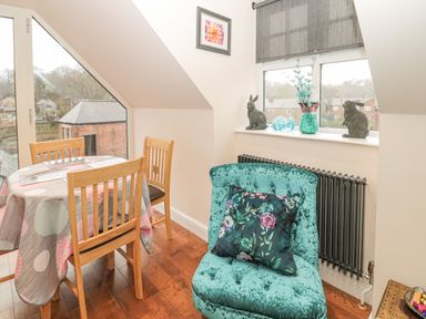 A dining area with wooden chairs and round table with tablecloth next to a window with rabbit figurines and a vase at River Cottage in Morpeth