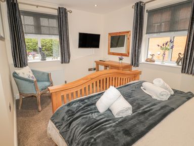 A bedroom with a wooden bed and folded towels on a gray blanket a blue chair a wooden desk a wall-mounted TV two windows with gray curtains and decorative items on the windowsills at River Cottage in Morpeth