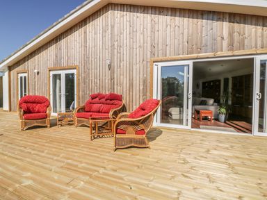 A wooden deck with wicker furniture and red cushions outside a wooden house with glass doors at Chy Lagen Hos in Newquay