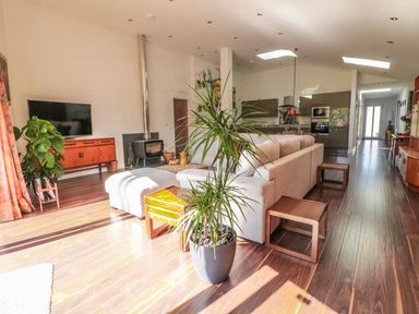 An open living room with a gray sectional sofa a wooden sideboard a flat screen tv a wood stove and a large potted plant at Chy Lagen Hos in Newquay