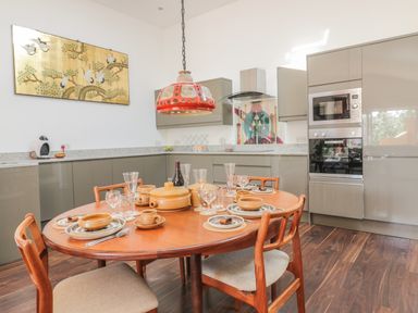 A kitchen with a round wooden dining table with dishware and glassware a hanging light and built in cabinets at Chy Lagen Hos in Newquay