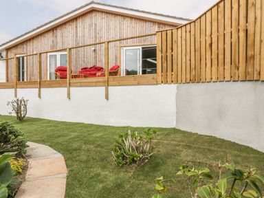 A garden with grass and shrubs outside a wooden house with a balcony and red cushions on chairs at Chy Lagen Hos in Newquay