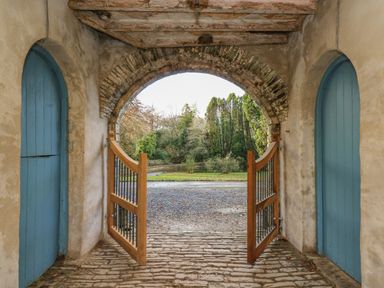 An arched stone passageway with wooden and metal gates opening to a gravel path and trees on either side blue wooden doors inside the arch at The Old Carriage Court in Kidwelly