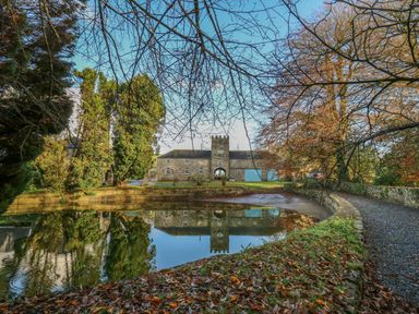 A stone building with blue doors reflected in a pond surrounded by trees and a gravel path at The Old Carriage Court in Kidwelly