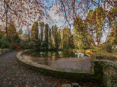 A circular stone pond surrounded by trees with autumn leaves on the ground at The Old Carriage Court in Kidwelly