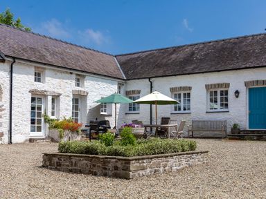 An outdoor courtyard with white stone buildings a raised flower bed a wooden table with umbrellas chairs and a bench at The Old Carriage Court in Kidwelly