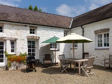 An outdoor patio area with a wooden dining table and chairs under umbrellas and a barbecue grill in front of a white stone building at The Old Carriage Court in Kidwelly