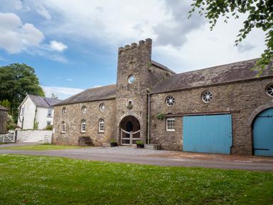 A stone building with blue doors and a tower surrounded by grass and a gravel driveway at The Old Carriage Court in Kidwelly