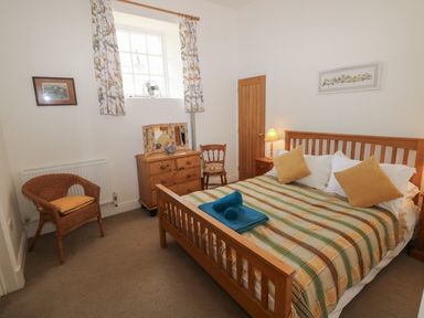A bedroom with a wooden bed with a checkered blanket and cushions a wooden dresser a wicker chair and a window with floral curtains at The Old Carriage Court in Kidwelly