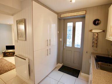An entrance area with a gray door with glass panels a black doormat white cabinets a kitchen sink and a wall clock with a fish decoration at Serendipity in Fowey