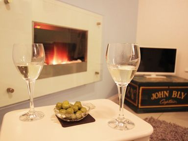 Two glasses of white wine and a bowl of green olives on a table near a fireplace and a television on a box labeled John Bly Captain