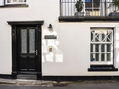 A black front door with glass panes next to a white wall with a window and a sign saying Serendipity at Serendipity in Fowey
