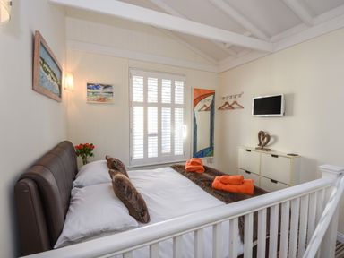 A bedroom with a bed white bedding orange towels a wall-mounted television and paintings at Serendipity in Fowey