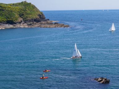 A coastal scene with kayakers and sailboats in the water near a rocky and green hillside at Serendipity in Fowey