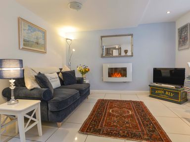 A living room with a dark sofa pillows a side table lamp an electric fireplace a patterned rug and a television at Serendipity in Fowey