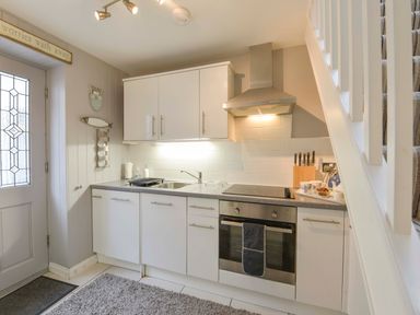 A kitchen with white cabinets an electric stove a stainless steel oven and a sink next to a door and a staircase at Serendipity in Fowey
