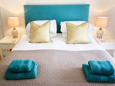 A bedroom with a double bed with two beige pillows bedside tables with lamps and folded blue towels at The Shell Seekers in Fowey