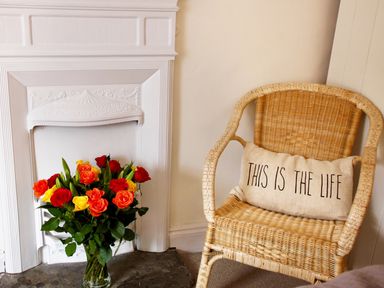 A wicker chair with a cushion that says this is the life next to a white fireplace with a vase of colorful roses at The Shell Seekers in Fowey