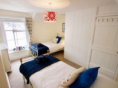 A small bedroom with two single beds blue blankets and pillows a window with patterned curtains and a white door with hooks at The Shell Seekers in Fowey