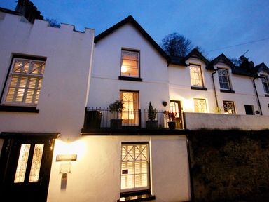Exterior view of a white two story house with lit windows and plants on the balcony at The Shell Seekers in Fowey