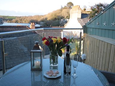 A balcony with a glass table holding a lantern with a candle flowers in a vase a bottle two glasses and a plate of snacks with rooftops and water in the background at The Shell Seekers in Fowey