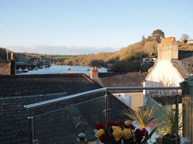 A balcony with flowers overlooking rooftops and a river with boats at The Shell Seekers in Fowey