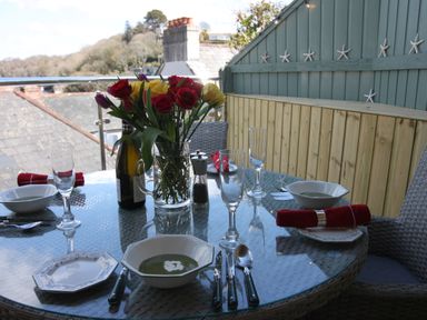 A glass outdoor dining table set with bowls of soup glassware cutlery and a vase of flowers on a balcony at The Shell Seekers in Fowey