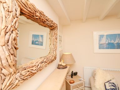 A corner of a room with a wooden framed mirror a small table lamp decorative bird figures framed sailboat pictures and a chair with pillows at The Shell Seekers in Fowey