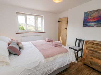 A bedroom with a bed with pink blankets and pillows a wooden dresser a black chair and a window overlooking the countryside at Cefn Nannau Ty-nant near Cerrigydrudion