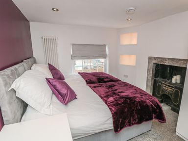 A bedroom with a double bed covered in white and purple bedding next to a window and a decorative fireplace at 11A Church Street Whitby