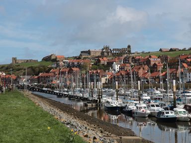 A marina with boats docked along the water and houses on a hillside with an abbey in the background at 11A Church Street in Whitby