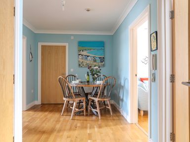 A dining area with a round table and four wooden chairs with a painting on the wall and a doorway to a living room at Tides Reach in Rhosneigr