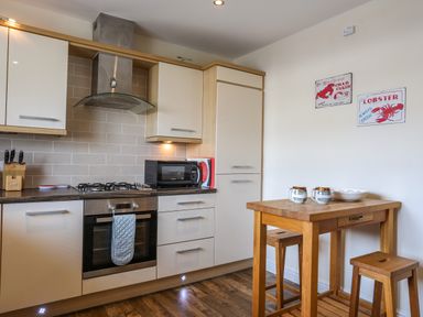 A kitchen with white cabinets a gas stove an oven a microwave a wooden table with two stools and two mugs on the table at Tides Reach in Rhosneigr
