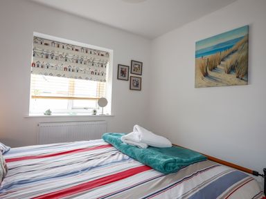 A bedroom with a striped bedspread folded towels on the bed a window with patterned blinds and paintings on the wall at Tides Reach in Rhosneigr