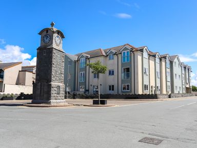 A clock tower on a paved area next to a three story residential building with multiple windows and balconies
