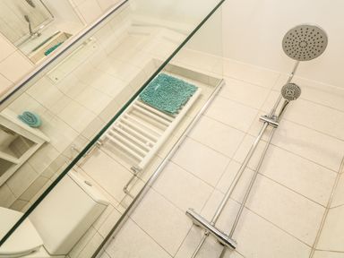 A bathroom with a shower area a glass shower screen a wall-mounted chrome shower head a white heated towel rail and a rolled turquoise towel at Gull Rock in Trebarwith Strand