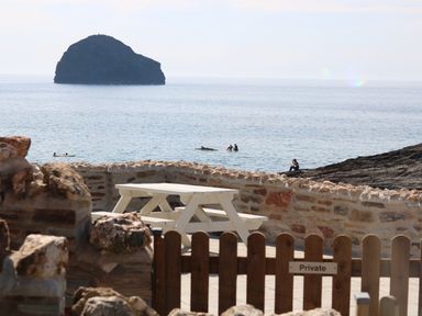 A stone patio with a white picnic table behind a wooden gate overlooking the sea with a large rock formation in the water at Gull Rock in Trebarwith Strand