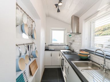 A narrow kitchen with hanging pots and pans a sink stove and window at Gull Rock in Trebarwith Strand