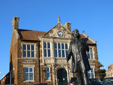 A statue of a man with a cane in front of a town hall building with a clock and tourist information signs in Hunstanton