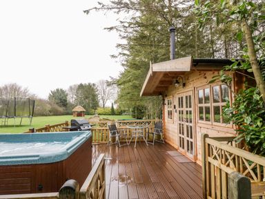 An outdoor wooden deck with a hot tub table with chairs and a barbecue grill next to a wooden cabin surrounded by trees at Someplace Else in Diss