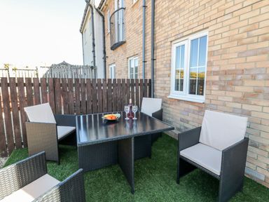 An outdoor patio area with four cushioned chairs around a square table with a bowl of fruit and wine glasses on an artificial grass surface at the beach house in beadnell