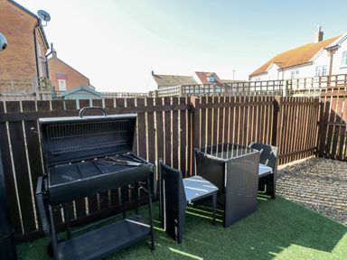 A fenced outdoor area with a barbecue grill and a small table with two chairs on artificial grass at Beach House in Beadnell