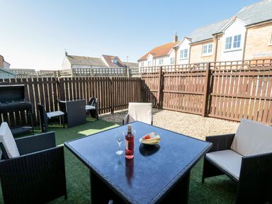 An outdoor patio area with a black table and chairs a bottle of wine and a fruit bowl on the table and houses in the background at Beach House in Beadnell